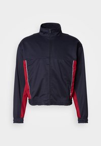 Ναυτικό μπλε μπουφάν με κόκκινες πλαϊνές πάνες, ψηλό κολάρο, μπροστινό φερμουάρ, ελαστικές μανσέτες και gathered στρίφωμα. Κατασκευασμένο από ομαλό, ελαφρύ ύφασμα.