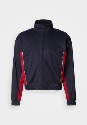 Navy blue jacket with red side panels, high collar, front zipper, elastic cuffs, and a gathered hem. Made from smooth, lightweight fabric.