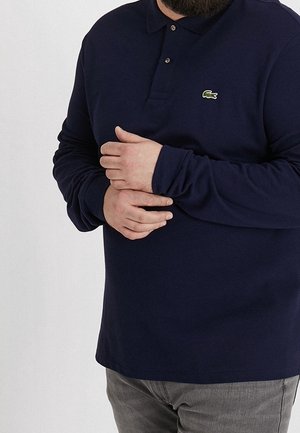 Osoba nosząca granatową koszulkę polo z długim rękawem z małym zielonym logo krokodyla, trzymająca lewe nadgarstek prawą ręką.