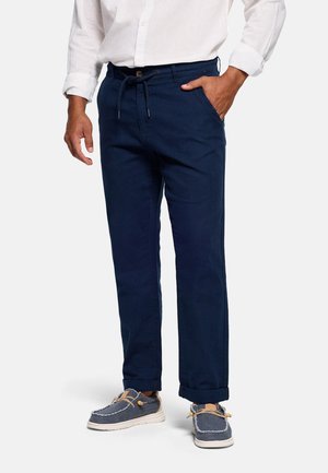 Hombre vistiendo pantalones con cordón azul marino, camisa blanca con mangas remangadas y zapatos casuales azul deslizables.