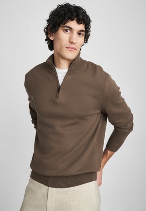 Hnedý pulóver na zips s textúrovaným pletením. Má rebrované manžety a spodný lem. Model ho nosí cez bielu košeľu s svetlo béžovými nohavicami.