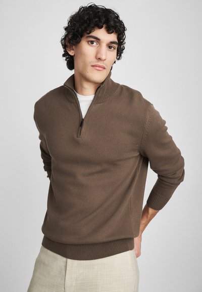 Pull à col zippé marron en tricot texturé. Présente des poignets et un ourlet côtelés. Modèle le portant par-dessus une chemise blanche avec un pantalon beige clair.