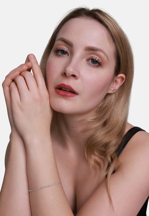 Nahaufnahme einer Person mit blonden Haaren, die ein schwarzes Oberteil trägt und ein zartes Silberarmband am Handgelenk zeigt, das kleine, klare Akzente aufweist.