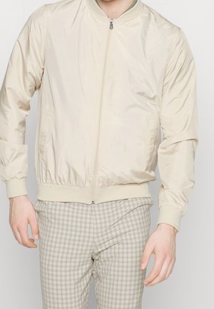 Homme portant une veste bomber beige clair zippée et un pantalon à carreaux gris clair, debout devant un fond blanc uni.