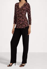 Kvinna klädd i en svart blommig wrap-top med 3/4 ärmar, svarta vida byxor och svarta högklackade sandaler, stående mot en vit bakgrund.