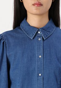 Camisa de mezclilla con cuello tipo solapa, mangas abullonadas y cierres de botones de perlas. La tela es de un azul intenso con una textura sutil.