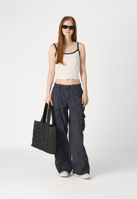 Melnas kvadrātveida rokassomas ar diviem rokturiem, valkāta ar bežīgu topu un tumši zilām platiem biksēm, ko papildina melnas brillēs.