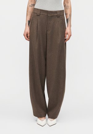 Brune bukser med vide ben båret af en tatoveret person med en grå ærmeløs top og hvide spidse sko, stående foran en ensfarvet baggrund.