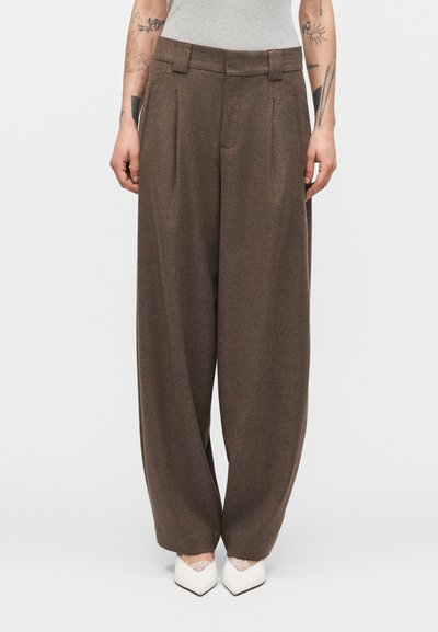 Wide-leg brown trousers worn by a tattooed person with a gray sleeveless top and white pointed shoes, standing against a plain background.