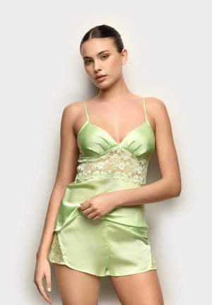 Junge Frau trägt ein hellgrünes Satin- und Spitzen-Camisole- und Shorts-Set und posiert vor einem neutralen, einfarbigen Hintergrund.