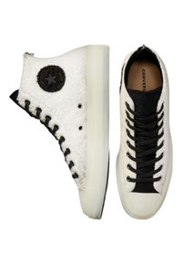 Höga och låga vita sneakers med svarta accenter, med en texturerad tygöverdela, runt svart logotyp och gummisulor.
