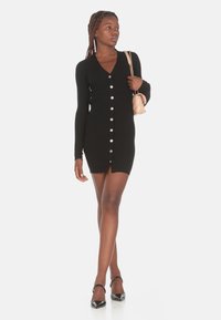 Abito nero a coste con maniche lunghe e scollatura a V, caratterizzato da una fila di bottoni argentati. Modella che tiene una piccola borsa beige, indossando tacchi neri.