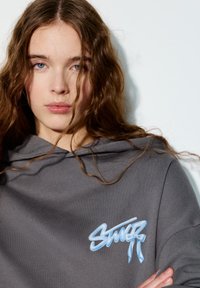 Siva majica s kapuljačom s plavim izvezenim logotipom s detaljem mašne. Tkanina ima glatku teksturu, a kapuljača dodaje ležerni štih.