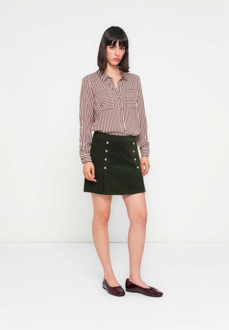 Chemise à rayures bordeaux et blanche avec deux poches poitrine, associée à une jupe en velours côtelé verte avec des boutons-pression en métal argenté et des chaussures plates.