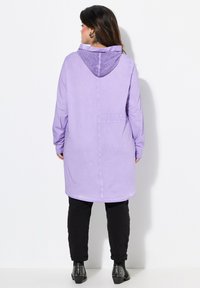 Paarse lange mouwen jas met een mesh capuchon, voorzien van een achterzak en een rechte snit, gedragen met zwarte taps toelopende broek en gestikte schoenen.