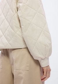 Μπεζ μπουφάν με καπιτονέ υφή από απαλό, ελαφρύ υλικό, με φουσκω τά μανίκια και κοντό σχέδιο. Συνδυάζεται με μπεζ παντελόνια.