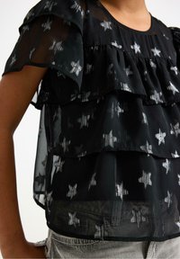 Schwarzes transparentes Oberteil mit silbernem Sternenmuster, mit geschichteten Rüschen und kurzen Ärmeln, aus leichtem Material gefertigt. Keine auffälligen Beschläge oder Akzente.