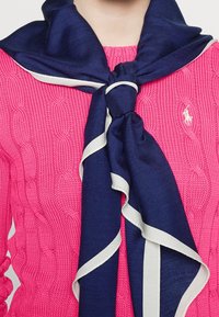 Bufanda azul marino con acentos crema, atada al cuello, sobre un suéter de punto trenzado rosa texturizado, exhibiendo una tela suave y colores contrastantes.