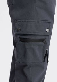 Tmavě šedé cargo kalhoty s kapsou na klopě, zabezpečenou dvěma kovovými patentkami, a kapsou na zip s černou poutkou na stehně.