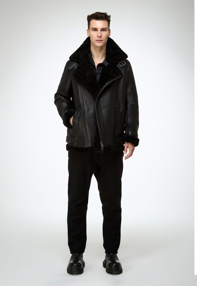 Schwarze Lederjacke mit großem schwarzem Lammfellkragen, Reißverschluss vorne und seitlichen Schnallen. Getragen mit schwarzer Hose und robusten schwarzen Stiefeln.