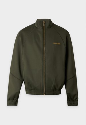 Homme portant une veste zippée vert foncé, un polo blanc, un pantalon cargo marron ample et des chaussures noires, debout sur un fond blanc.