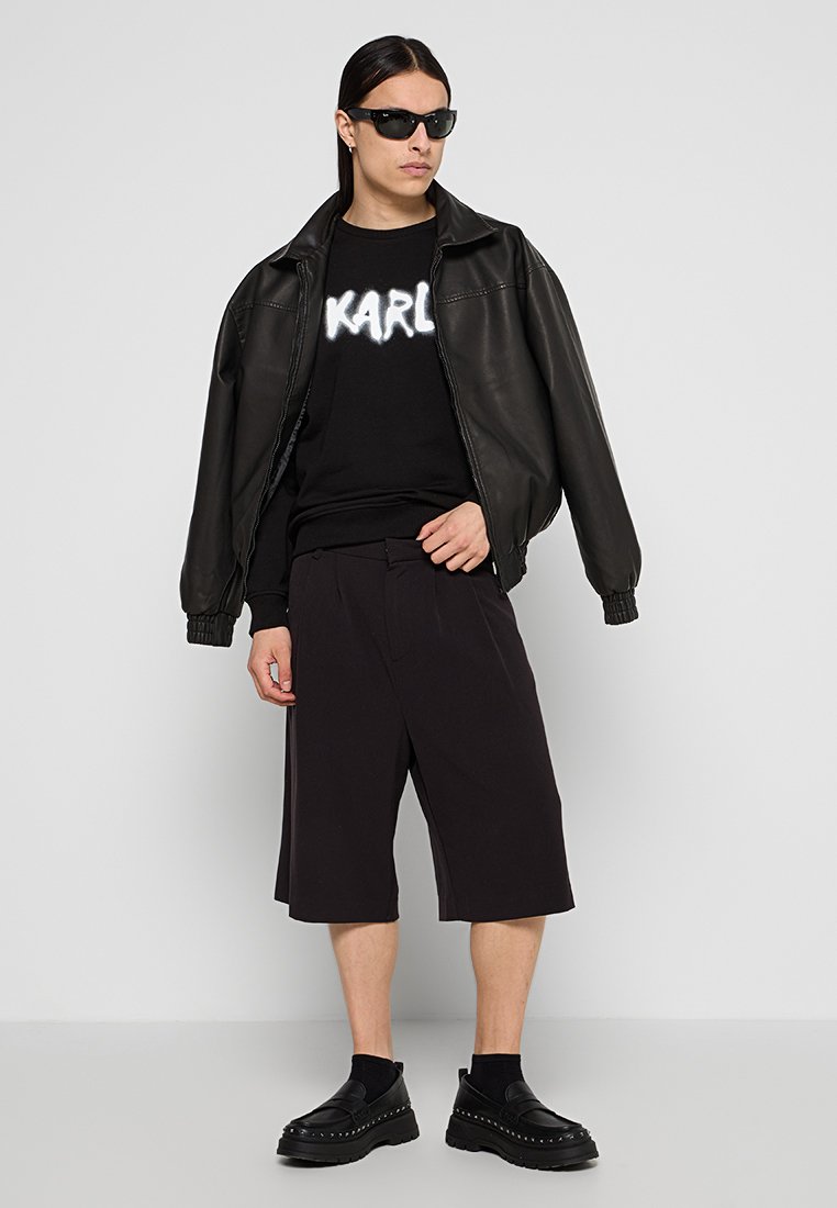 Chaqueta bomber de cuero negro, sudadera negra con texto blanco "KARL", pantalones cortos negros a la altura de la rodilla y zapatos slip-on negros gruesos.