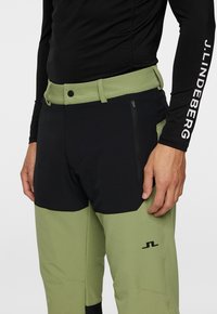 Mustat ja oliivinvihreät hiihtohousut, joissa on vetoketjullinen tasku, vyö ja logo alhaalla lahkeessa. Teksturoitu kangas ja voucher-mallinen muotoilu.