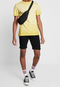 Camiseta amarilla a rayas, pantalones cortos negros, riñonera negra, calcetines blancos con rayas rojas y azules, zapatillas negras. Atuendo sencillo y casual.