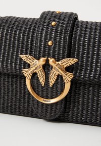 Zwarte geweven handtas met gouden vogelvormige sluiting en gouden studs op de band, die gedetailleerde textuur en decoratieve hardware toont.