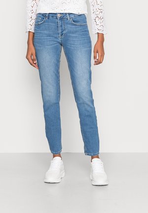 Person iført lyseblå slim-fit jeans, hvide sneakers og en hvid langærmet top med hulmønsterdetaljer, stående foran en ensfarvet baggrund.