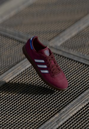 Bordowe buty sportowe z trzema białymi paskami, brązowymi sznurówkami i niebieskimi akcentami. Zamszowa tekstura z gumową podeszwą, lekko uniesioną cholewką.