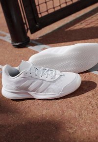 Coppia di scarpe sportive bianche con suole strutturate e tre strisce laterali posate su un campo esterno rossastro vicino a una recinzione metallica nera.