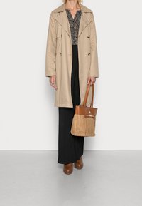 Beiger Trenchcoat über einem gemusterten Oberteil, schwarzen, weit geschnittenen Hosen und braunen High-Heel-Clogs. Geflochtener, sandfarbener Beutel mit braunem Griff.