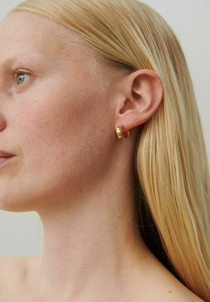 Blondīne ar gaišu ādu, nēsājot mazu zelta stīpu auskaru, redzama tuvplānā profilā pret vienkāršu fonu.