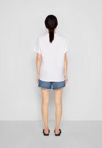 Žena s tmavými vlasy v culíku, oblečená v bílém tričku, roztrhaných denimových šortkách a černých sandálech, stojí čelem ke šedé zdi.