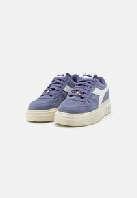 Sneaker in suede viola con un accento laterale in pelle bianca, lacci piatti e una suola beige spessa. Presenta perforazioni sulla punta.