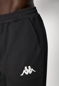 Melni sporta bikses ar maigu tekstūru, elastīgu jostu un balto logotipu uz kabatas. Dizainā ir iekļauta sānu kabata.