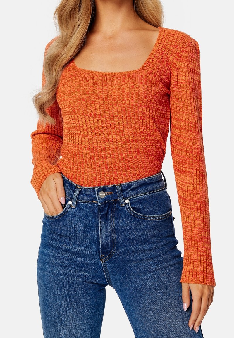 Femme portant un haut orange texturé à manches longues avec un décolleté carré et un jean bleu taille haute, main dans la poche.