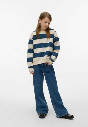 Jeune femme portant un pull rayé bleu et blanc, un jean à jambes larges bleu et des chaussures noires, debout avec une main dans la poche.