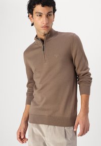 Sweater zippé marron en tissu tricoté doux. Présente un col zippé à moitié, des poignets et un ourlet côtelés. Logo de la marque discrètement brodé sur la poitrine.
