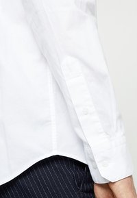 Witte katoenen overhemdsmouw met een puntige manchet en knoopsluiting. Heeft een subtiele textuur, afgebeeld naast gestreepte broek.