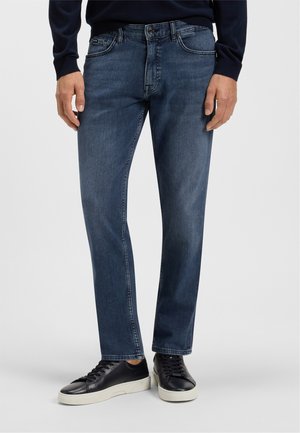 Donkerblauwe denim jeans met een rechte snit, vijf zakken en een ritssluiting. Draagt een paar zwarte leren sneakers met een witte zool.
