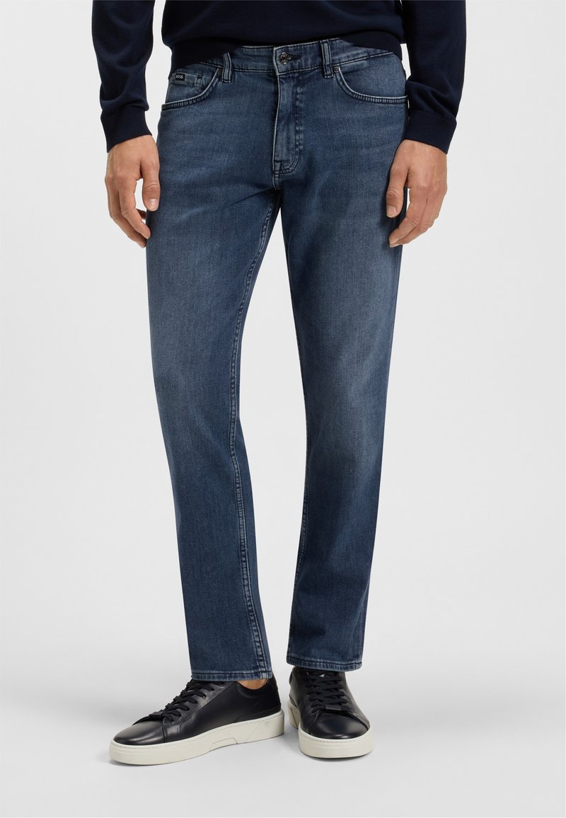 Dark blue denim jeans with a straight cut, five pockets, and a zip fly. Worn with black leather sneakers featuring a white sole.