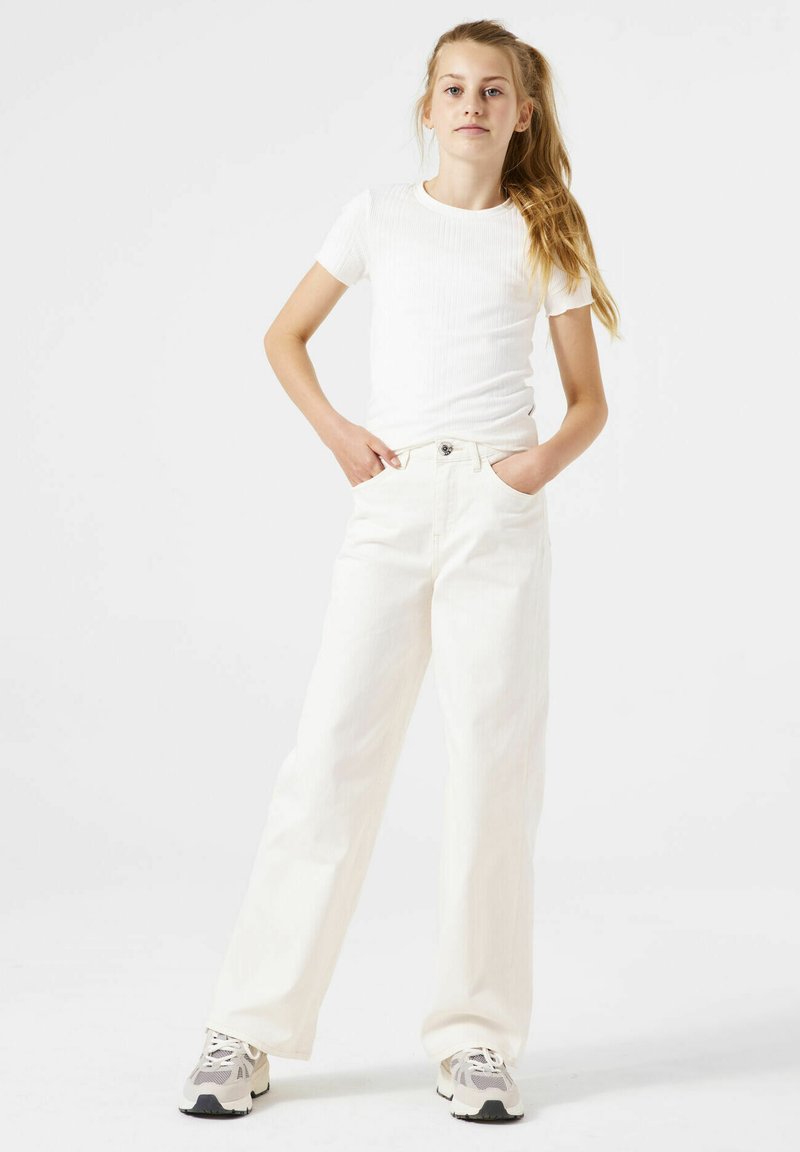 Camiseta blanca de manga corta con textura acanalada, combinada con pantalones de mezclilla blancos de pierna ancha y cintura alta. El modelo está de pie con las manos en los bolsillos frontales, luciendo zapatillas.