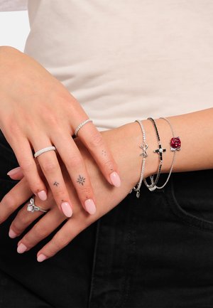 Hands with pale pink nails wearing silver rings and three silver bracelets featuring a bow, cross, and red rose charm on a light skin tone.
