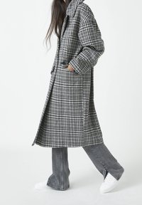 Harmaa ruudullinen pitkä takki, jossa kaulus, kaksi sivutaskua ja väljähkö istuvuus, yhdistetty harmaisiin levenevillä lahkeilla varustettuihin farkkuihin ja valkoisiin tennareihin. Sileä kangaspinta.