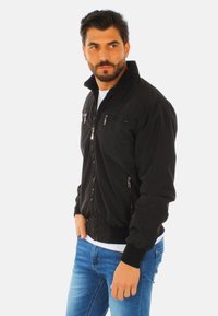 Giubbotto bomber nero con tasche con zip, polsini e colletto a coste. Indossato sopra una camicia bianca e jeans blu, che mettono in evidenza un design aderente.