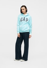 Jeune personne debout, portant un sweat à capuche bleu clair de la marque GAP, un jean large et sombre, ainsi que des baskets noires et blanches, sur un fond uni.
