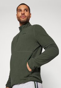Pullover in pile verde con colletto a mezza zip, tasche laterali e un logo discreto sul petto. Texture morbida con una silhouette aderente.