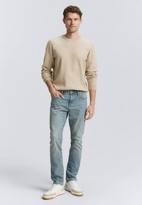 Béžový pletený svetr, světle modré úzké džíny a bílé tenisky s hnědými detaily. Model stojí, ruce v kapsách, na jednoduchém pozadí.